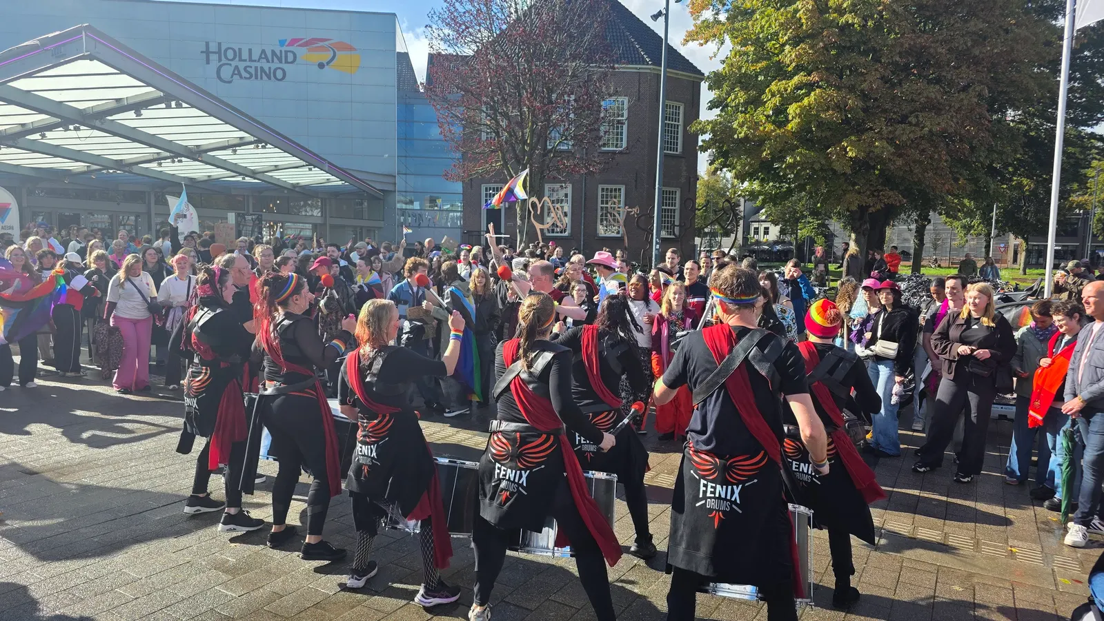 Fenix Drums speelt voor een grote menigte op een stadsplein, met regenboogvlaggen op de achtergrond